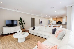 A modern living room with a flat-screen TV, white sofas, a coffee table, and a kitchen in the background. - Lumina A Spacious Haven Close to Adelaides Best (Allenby Gardens)
