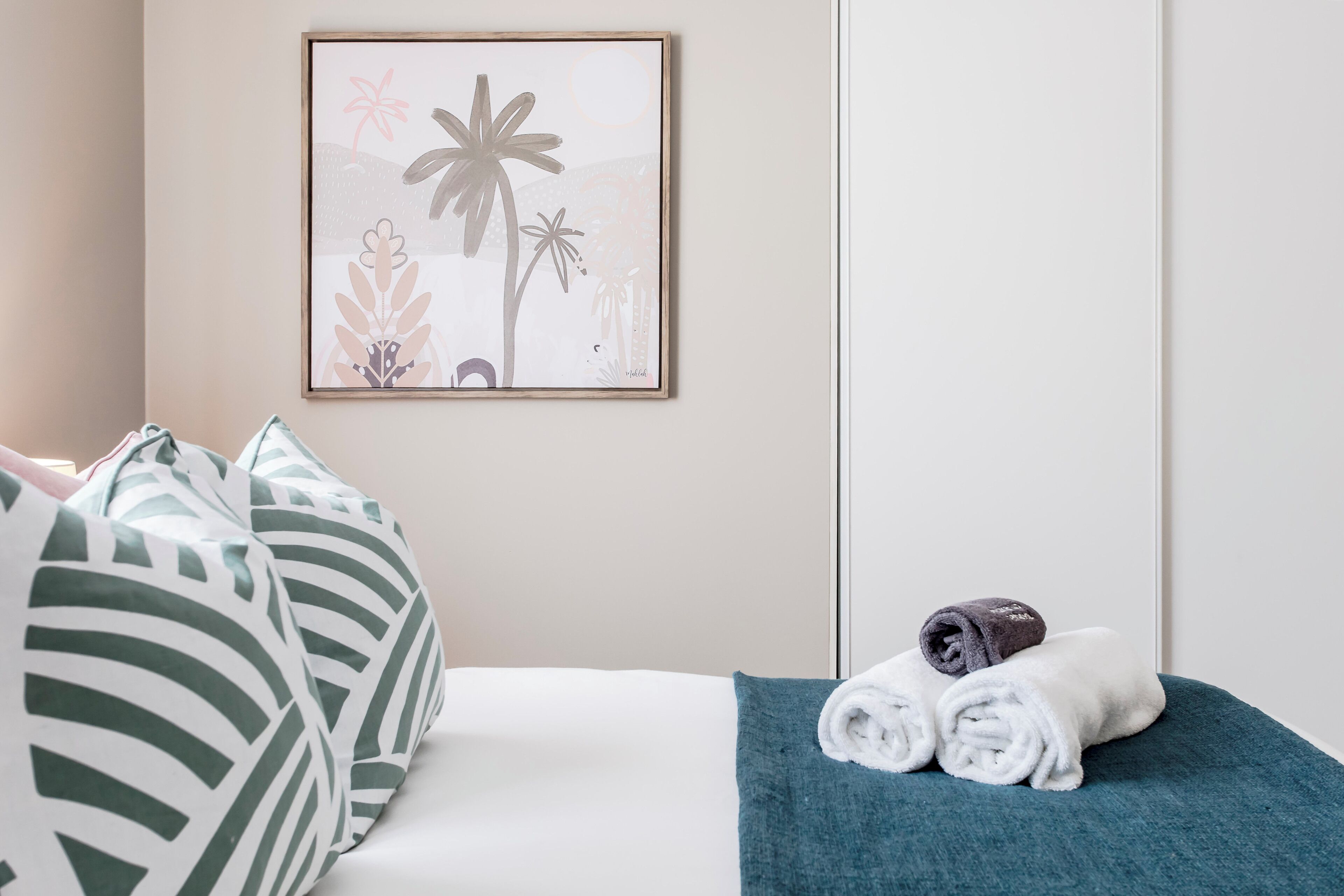 A neatly made bed with a teal blanket, a rolled towel, and a framed artwork of tropical plants on the wall.