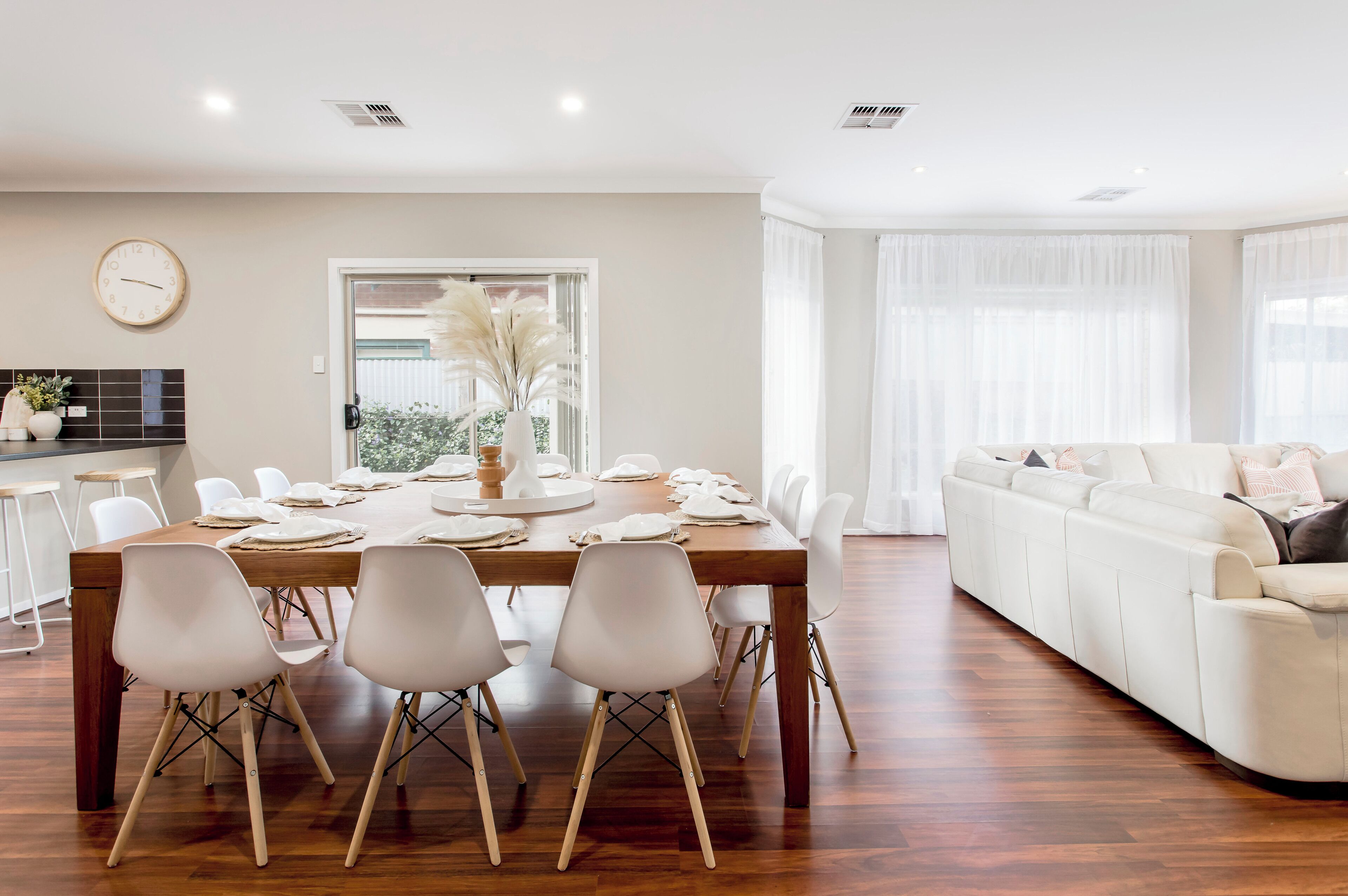 A dining area with a wooden table, white chairs, a clock on the wall, and a white sofa in the background.