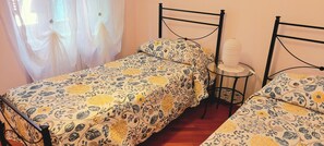 Ein Zimmer mit zwei Einzelbetten, Blumenmuster auf der Bettwäsche, einem kleinen Tisch mit Lampe und einem Fenster mit durchsichtigen Vorhängen.