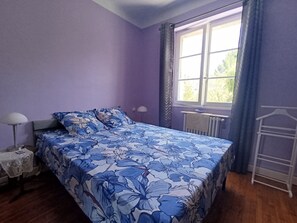Un dormitorio con cama, mesita de noche, lámpara, radiador y ventana con cortinas.