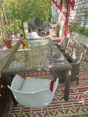 Una mesa con cubierta de vidrio rodeada de sillas de mimbre y un banco, con plantas en macetas y una pared de piedra al fondo.