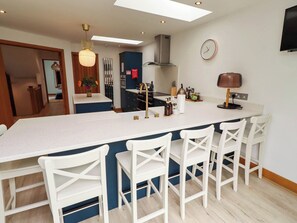 A modern kitchen with a large island, white bar stools, a clock, and a TV mounted on the wall. - Alma House (Seahouses)
