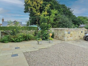 A stone wall with a sign indicating a parking area, a gravel driveway, and a variety of greenery including a large tree and potted plants. - Alma House (Seahouses)