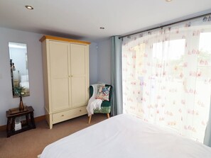 A bedroom with a large bed, a wardrobe, a mirror, a small table with a vase, and a window with curtains. - Alma House (Seahouses)