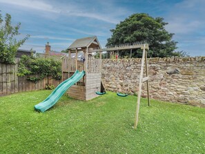 A wooden play structure with a slide and swings, set against a stone wall and a wooden fence. - Alma House (Seahouses)