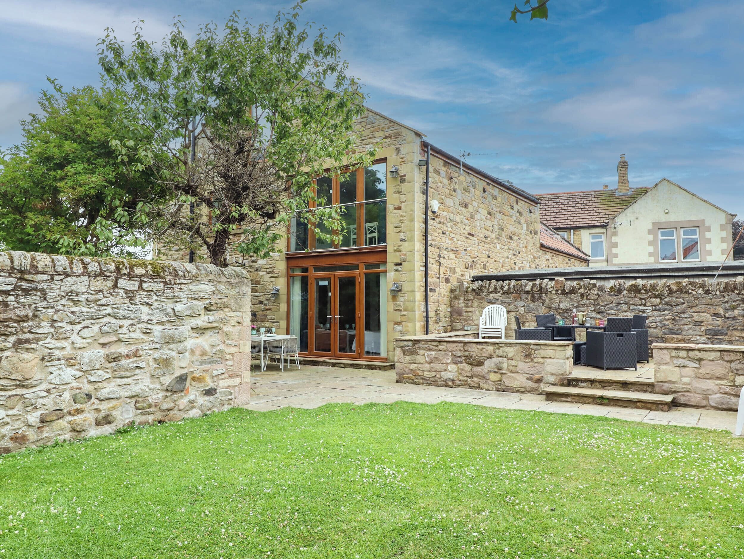 A stone building with a wooden door and windows, a stone wall, and a grassy yard.