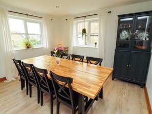 A dining room with a wooden table, black chairs, a vase with flowers, and a black cabinet with glass doors. - Alma House (Seahouses)