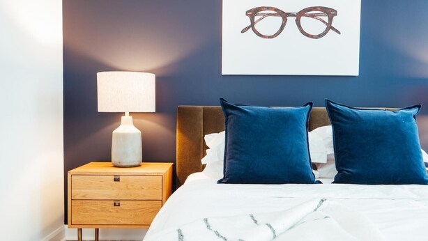 A bedroom with a bed, two pillows, a nightstand with a lamp, and a wall-mounted picture of glasses. - Landing Amazing Studio (Orlando)