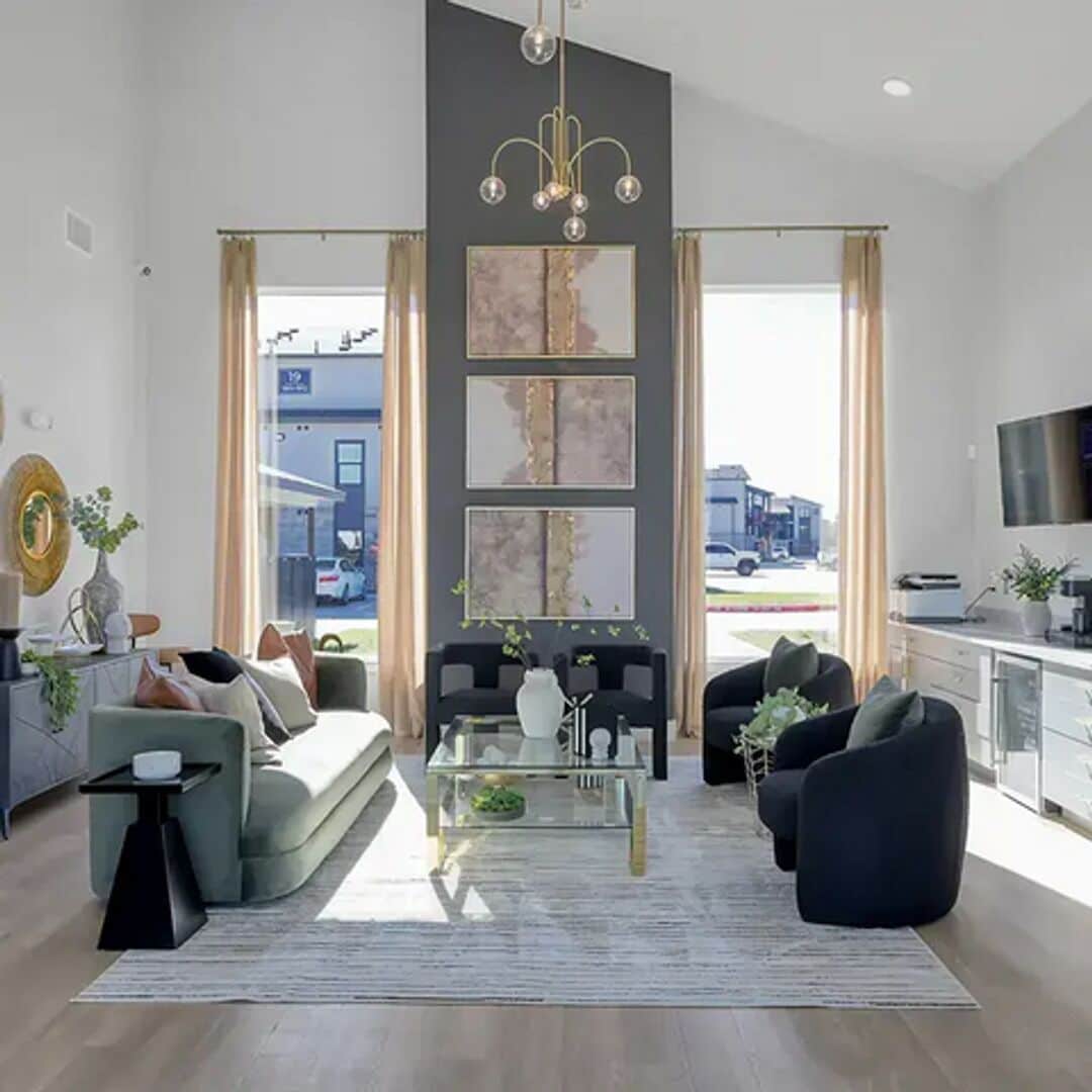 A modern living room with a sofa, armchairs, a coffee table, and a TV. There are large windows with curtains, a chandelier, and artwork on the wall.