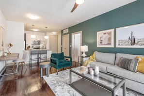 A modern living room with a grey sofa, teal armchair, and a glass coffee table. There is a kitchen area with white cabinets and a dining set. - Landing Amazing 1BD in Bear Creek (Houston)