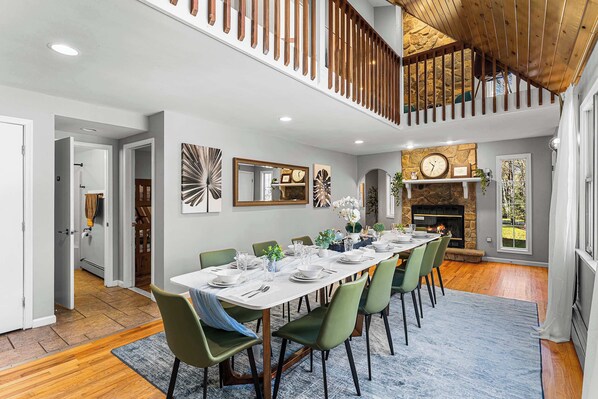 A dining area with a long table set for six, surrounded by green chairs, a stone fireplace, and a view of a garden through a window.