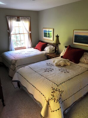 Ein Schlafzimmer mit zwei Betten, einem fensterartigen Durchbruch mit Vorhängen, einer Lampe und zwei gerahmten Bildern an der Wand.