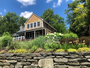 Ein zweistöckiges Haus mit einer Mauer aus Stein im Vordergrund, einem Holzdach und einem Garten mit verschiedenen Pflanzen und Blumen.