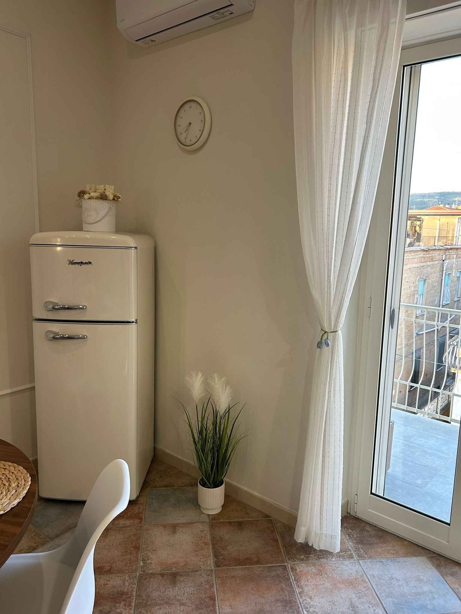 A compact room with a white refrigerator, a clock on the wall, a potted plant, and a view through a window with curtains.
