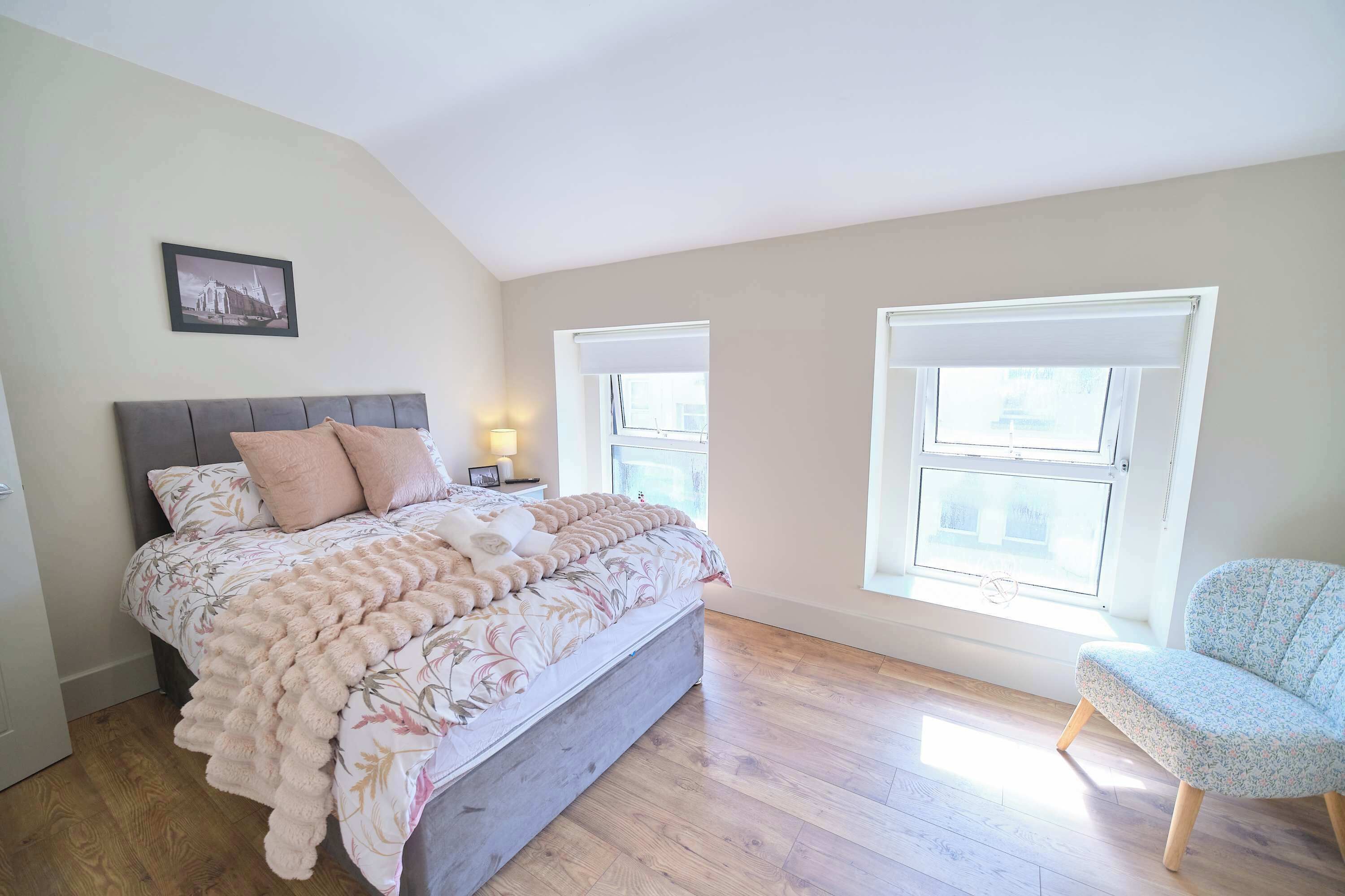 A bedroom with a bed, a chair, a nightstand, and two windows.