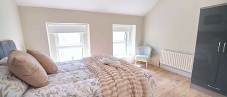 A bedroom with a bed, two windows, a chair, a radiator, and a wardrobe.