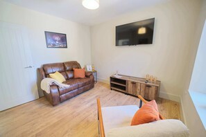 Ein Wohnzimmer mit braunem Ledersofa, einem Holztisch, einem an der Wand montierten Fernseher und einer Tür.