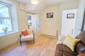 Ein Zimmer mit Holzboden, einem weißen Stuhl mit orangefarbener Sitzkissen, einem braunen Ledersofa mit gelber Sitzkissen, einem weißen Heizkörper und zwei gerahmten Bildern an der Wand.