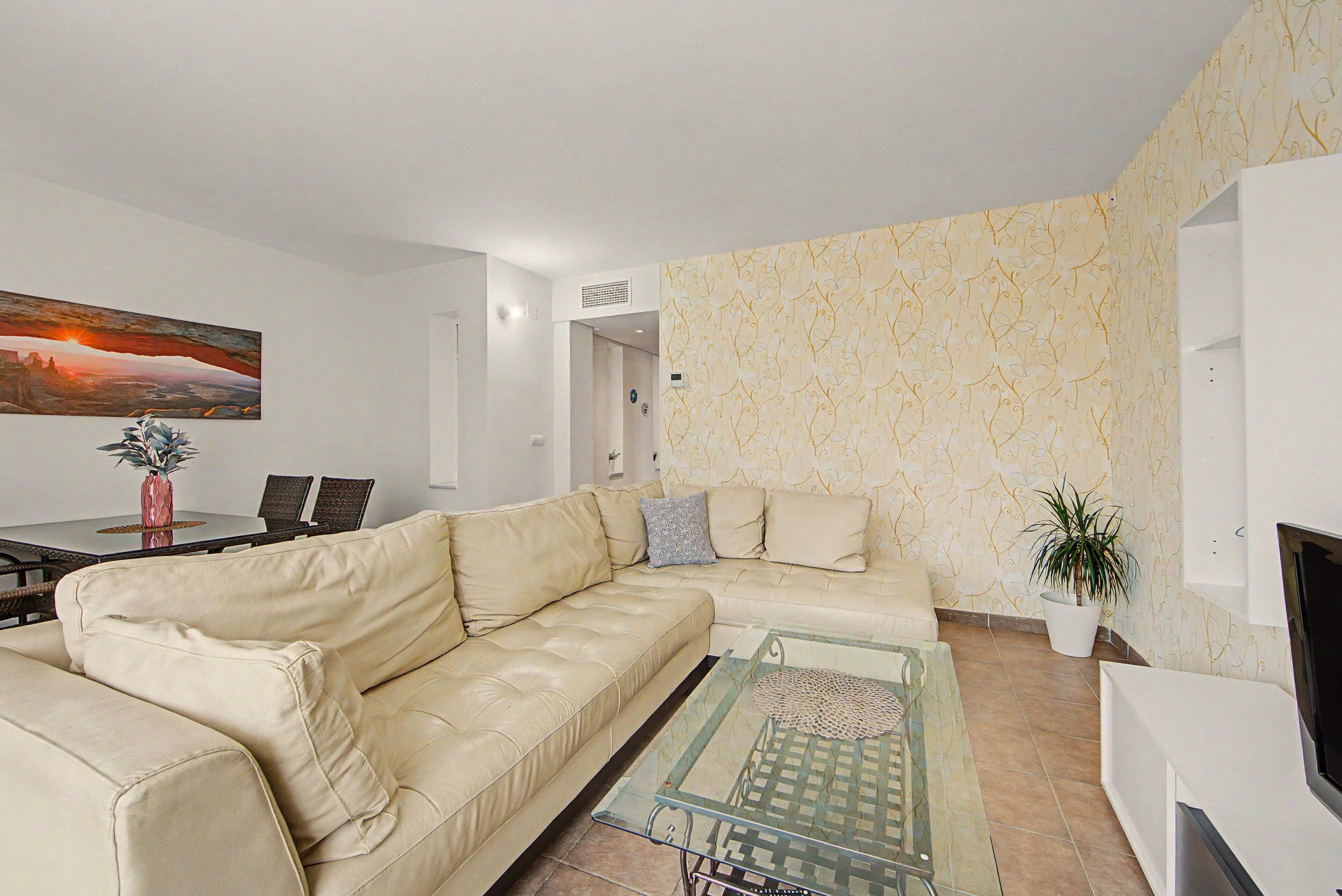 A living room with a beige sofa, a glass coffee table, a dining table with chairs, and a patterned wallpaper.