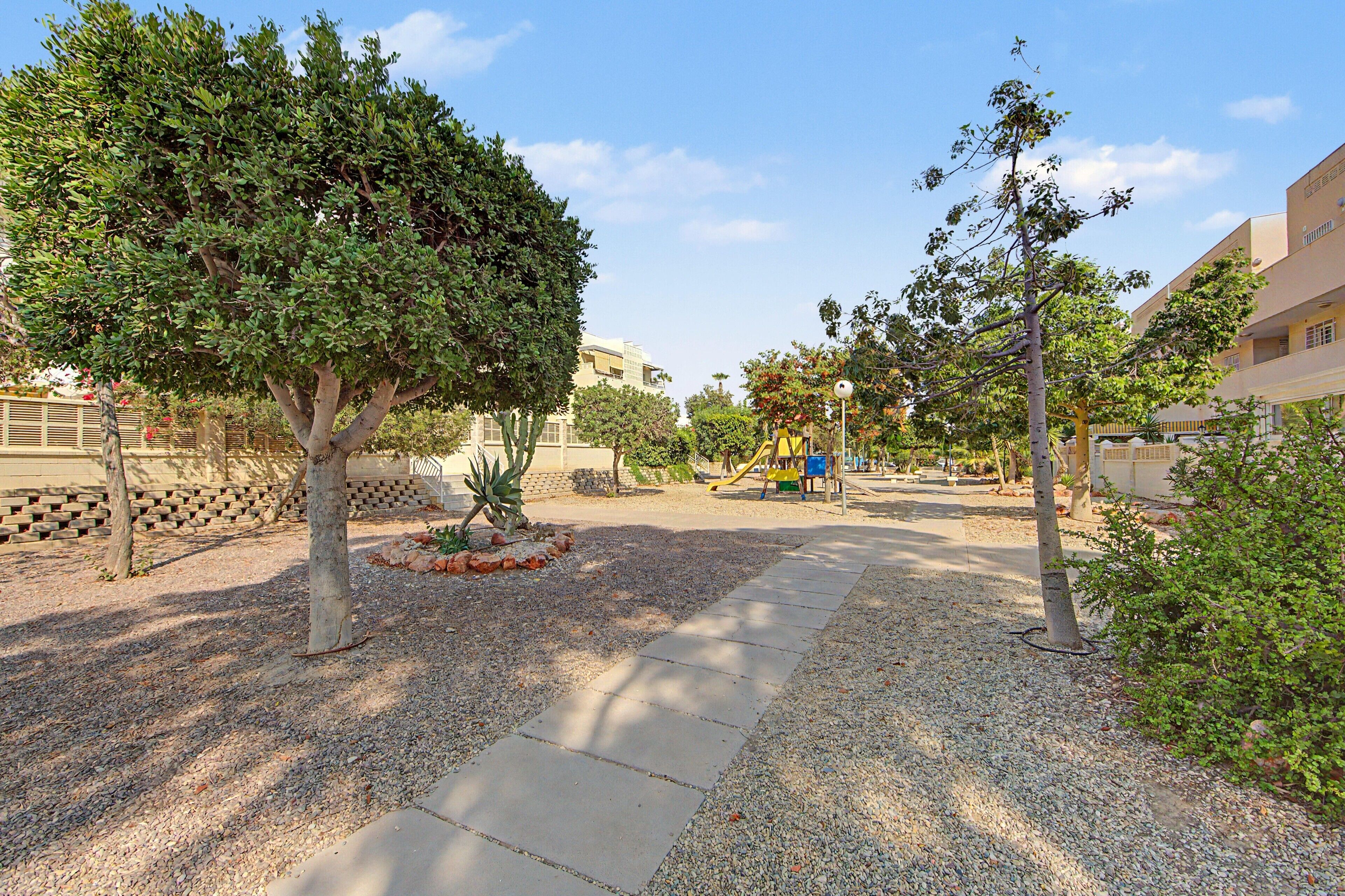 A paved walkway with gravel borders, surrounded by trees and a playground.
