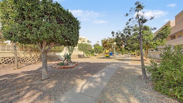 A paved walkway with gravel borders, surrounded by trees and a playground.