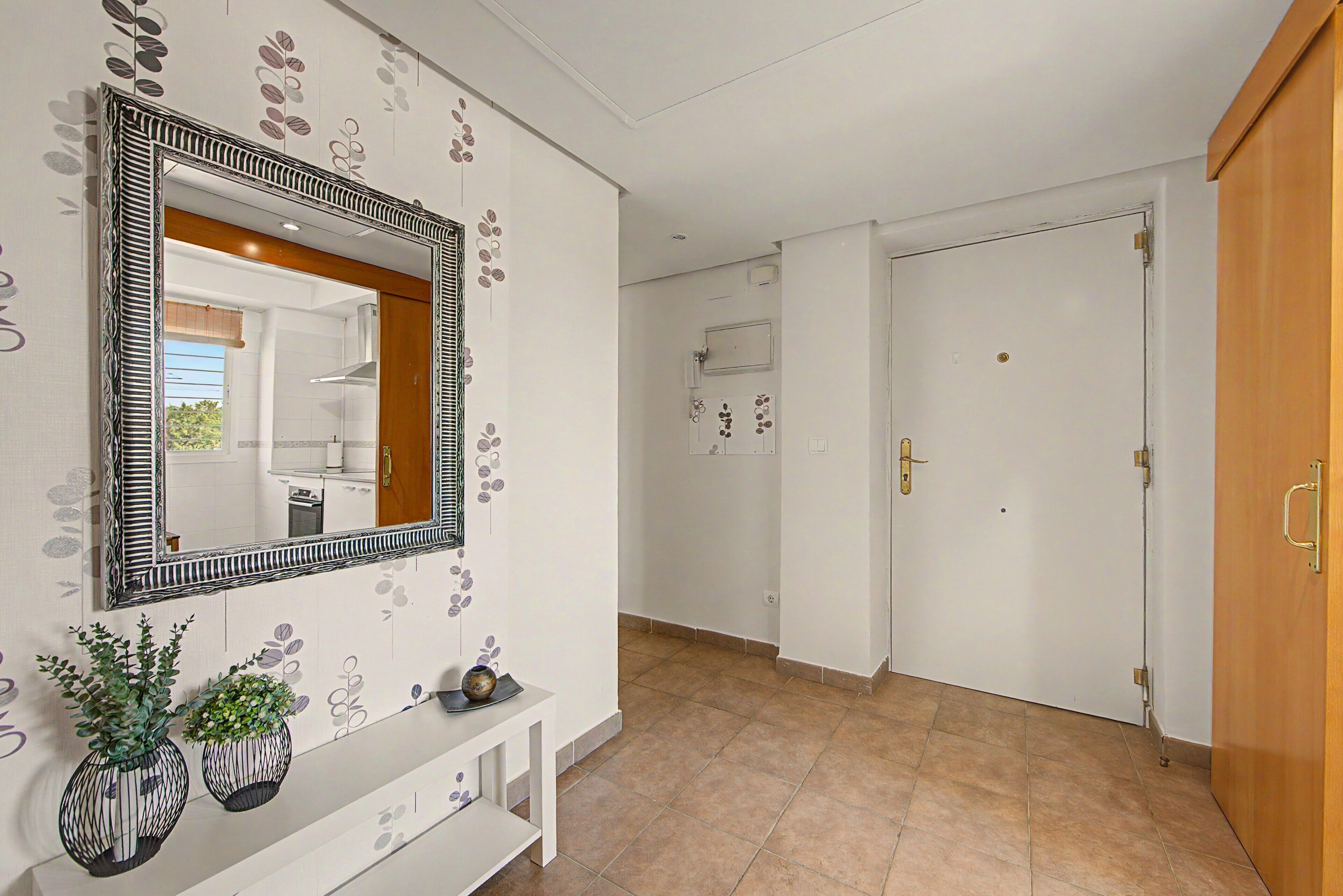 A hallway with a white door, a mirror with a decorative frame, and a console table with two potted plants.