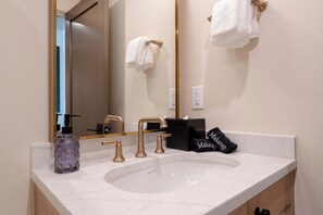 Une salle de bain avec un lavabo blanc, des robinets dorés, un grand miroir et un porte-serviettes sur lequel est posée une serviette blanche.
