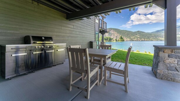 Een terras buiten met een barbecue, eettafel en stoelen, met uitzicht op een meer en bergen.