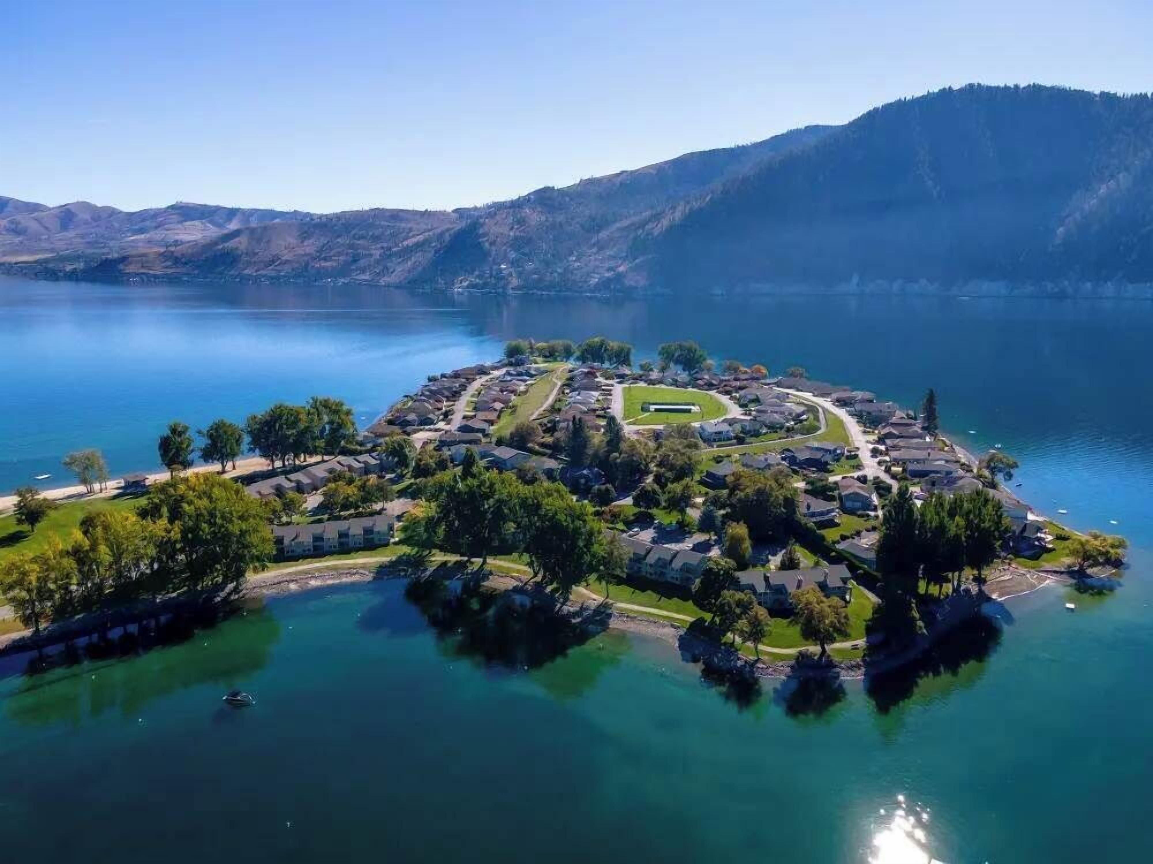 Vue aérienne d’un quartier résidentiel au bord d’un lac, avec des maisons, un espace vert central et des collines environnantes.