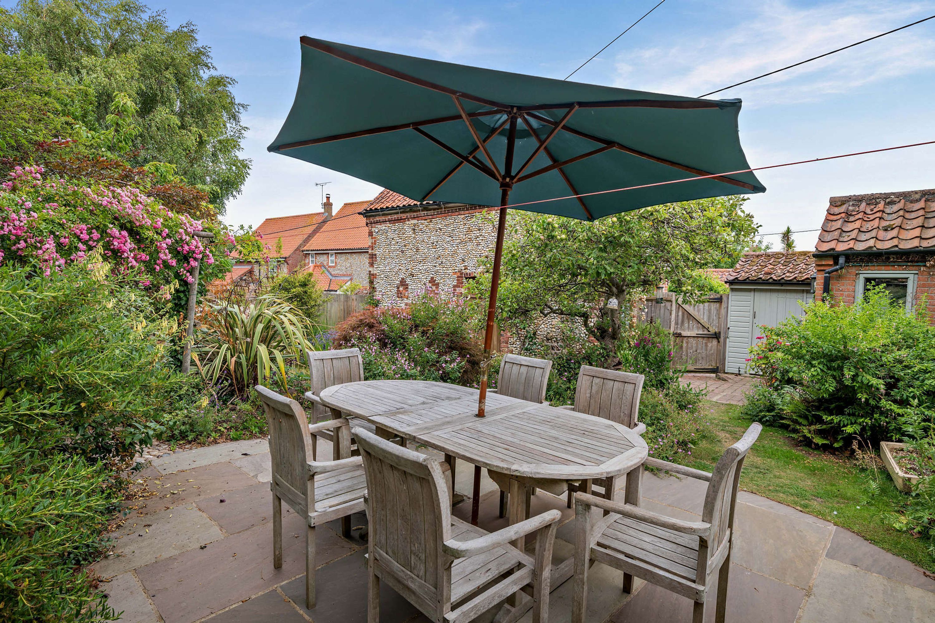 Een terras buiten met een houten tafel en stoelen, een groene parasol en een pad van stenen.