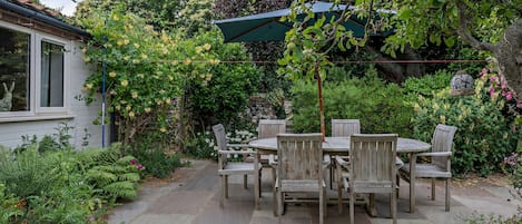 A patio with a table and chairs, surrounded by lush greenery and a stone pathway.