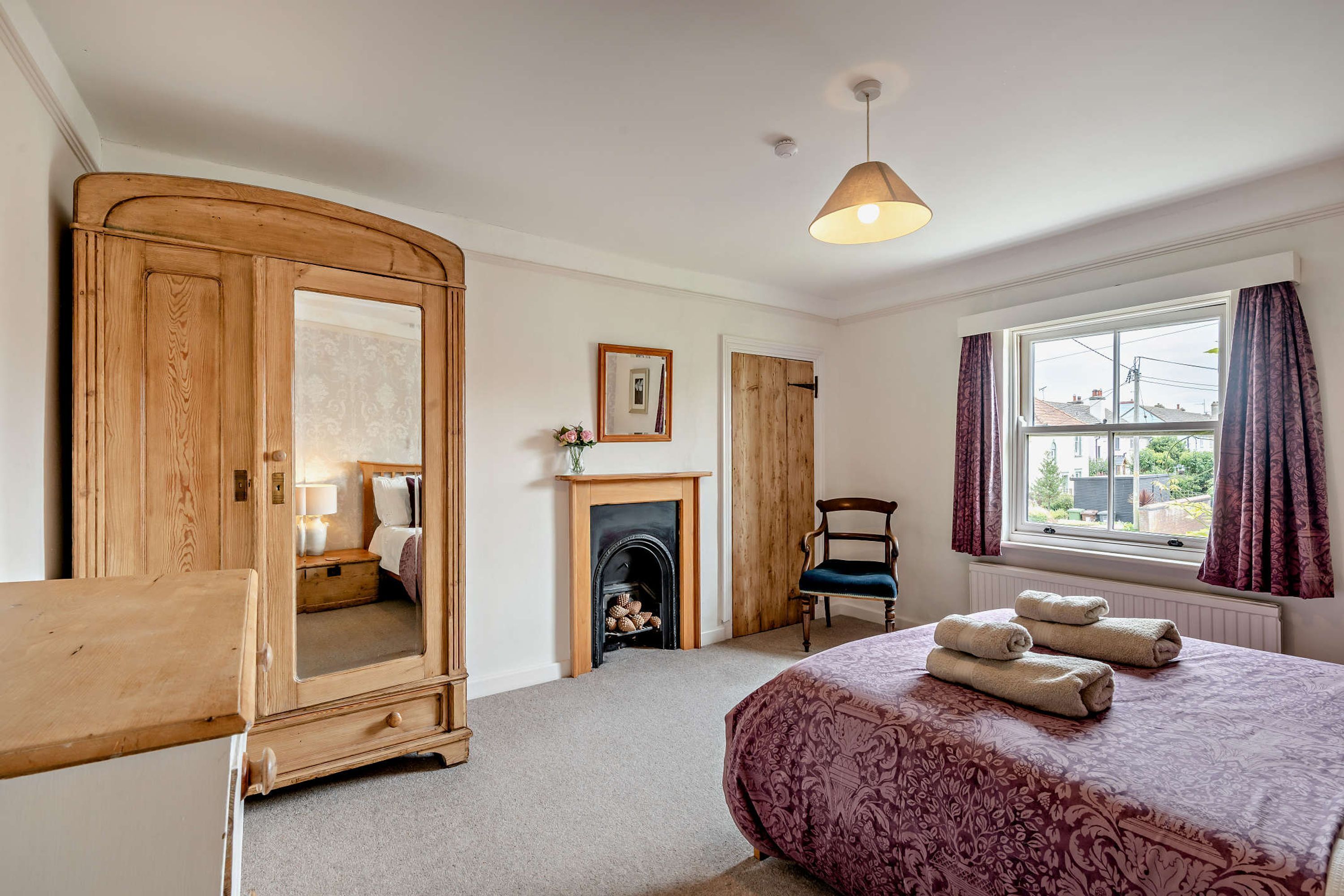 A bedroom with a bed, a wooden wardrobe, a fireplace, a chair, and a window with curtains.