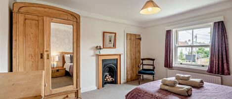 A bedroom with a bed, a wooden wardrobe, a fireplace, a chair, and a window with curtains.