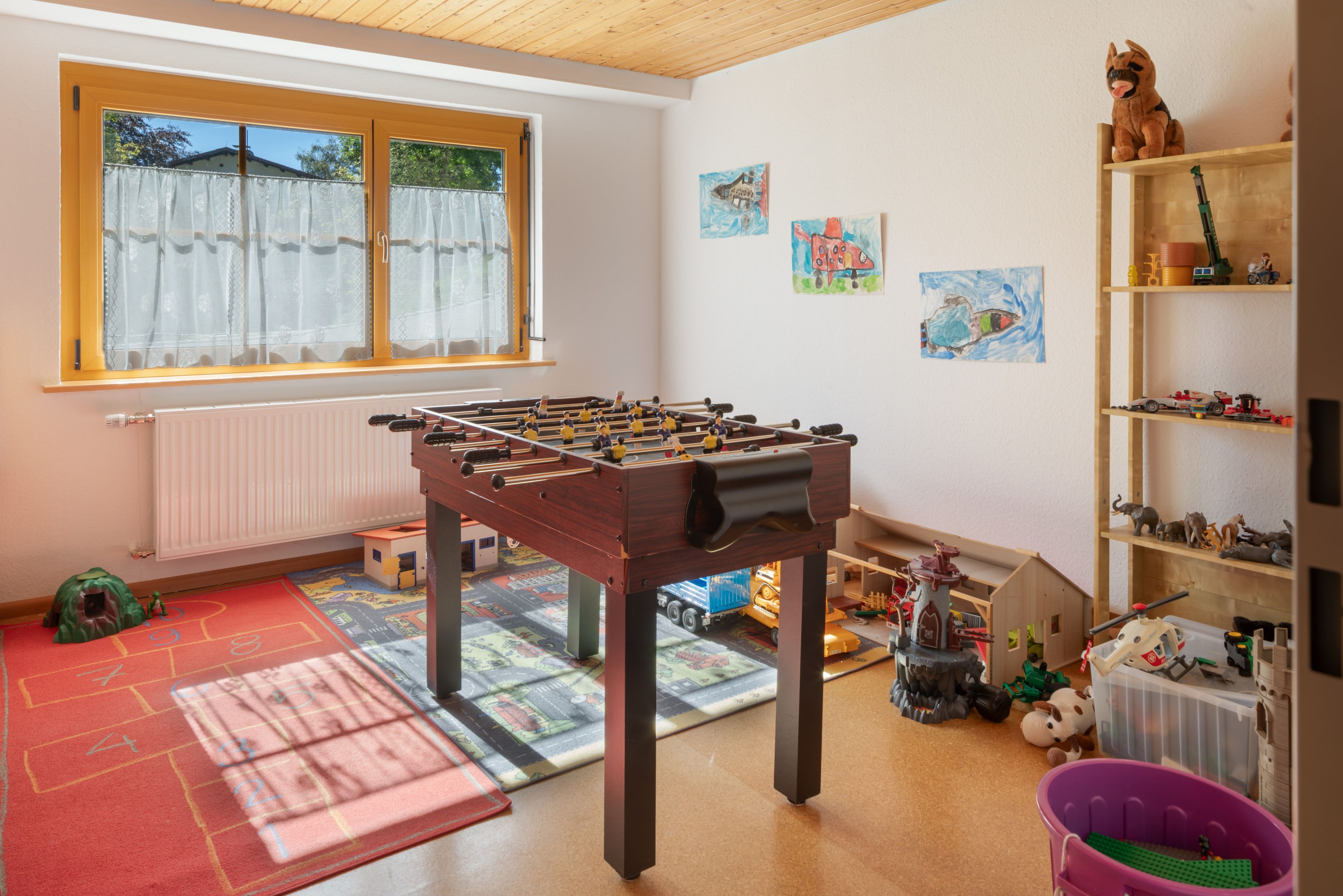 Et værelse med et fodboldbord, et vindue med gardiner, en radiator, et legeområde med måtte og en reol med legetøj.