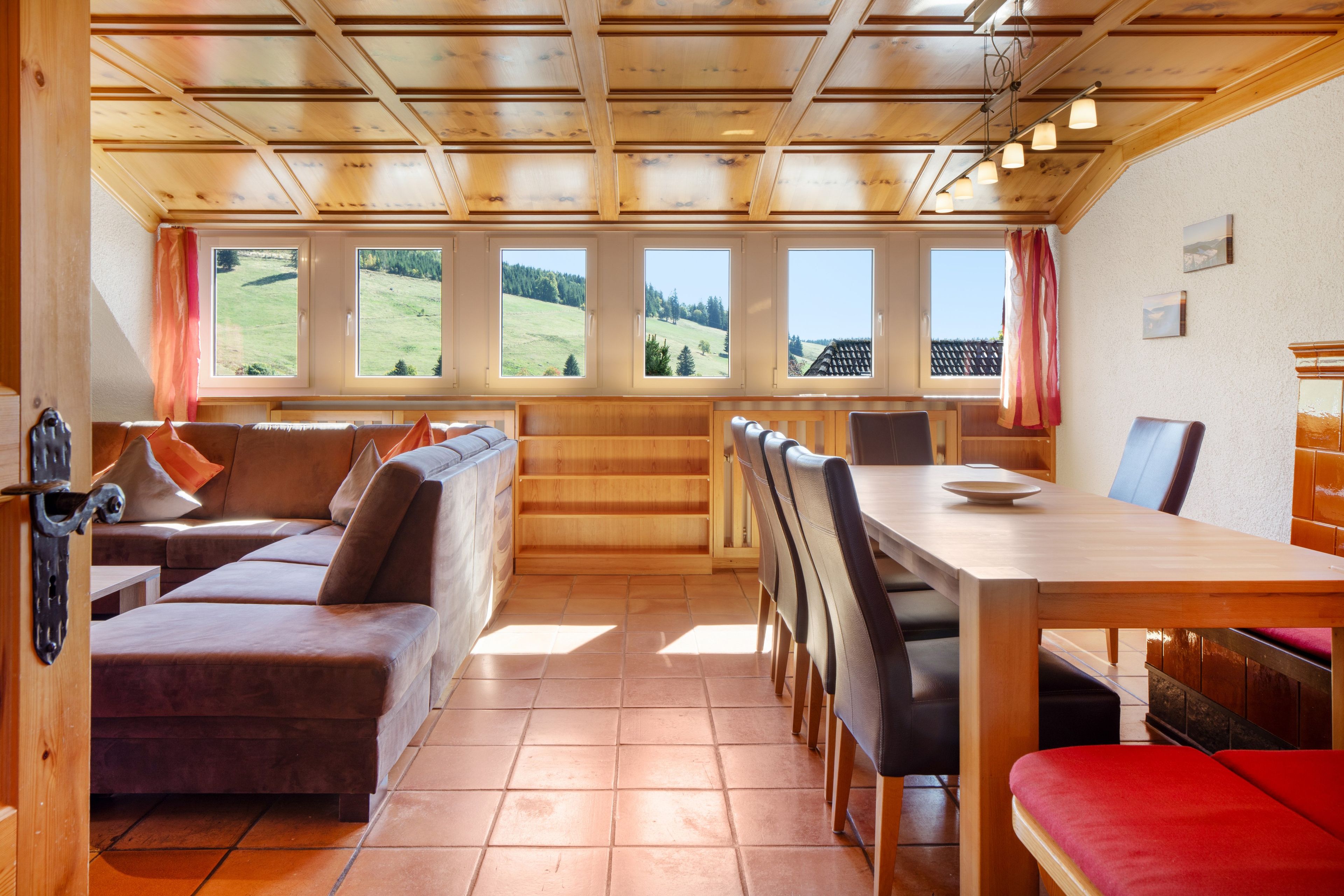 A cozy room with a wooden ceiling, a sofa, a dining table, and a view of a green landscape through large windows.