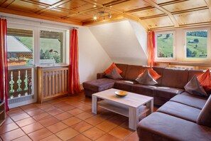 Ein gemütliches Wohnzimmer mit einer braunen Couch, einem Holztisch und Blick auf eine grüne Landschaft durch die Fenster.