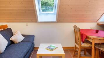 Een kamer met een houten plafond, een tafeltje met een boek, een bank met kussens en een eettafel met een rode tafelkleed.