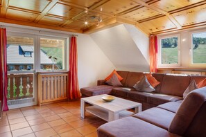 Een gezellige woonkamer met een bruine leren bank, een houten bijzettafeltje en uitzicht op een huis door een groot raam.