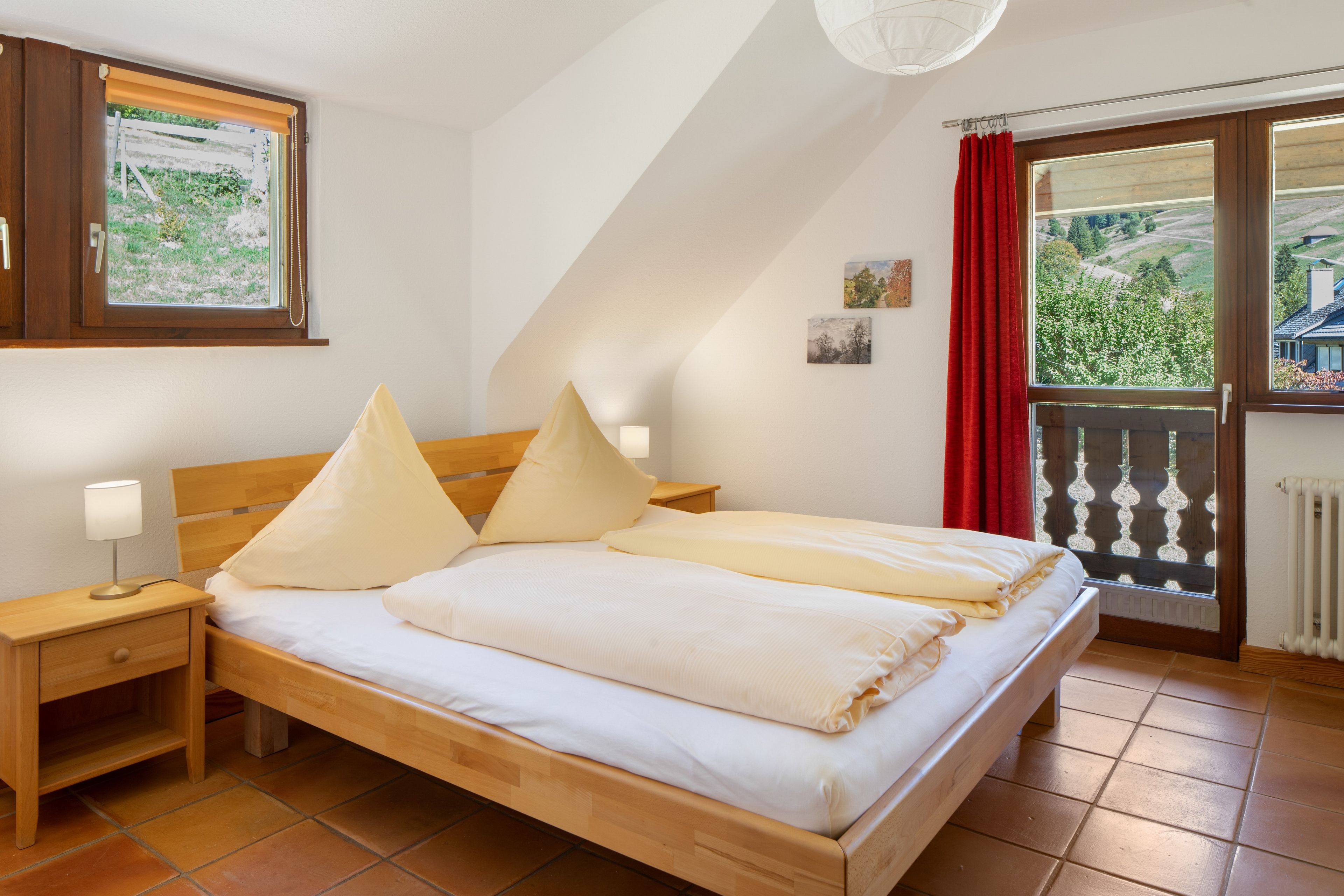 A bedroom with a wooden bed, a nightstand, a lamp, a window with a view of greenery, and a red curtain.