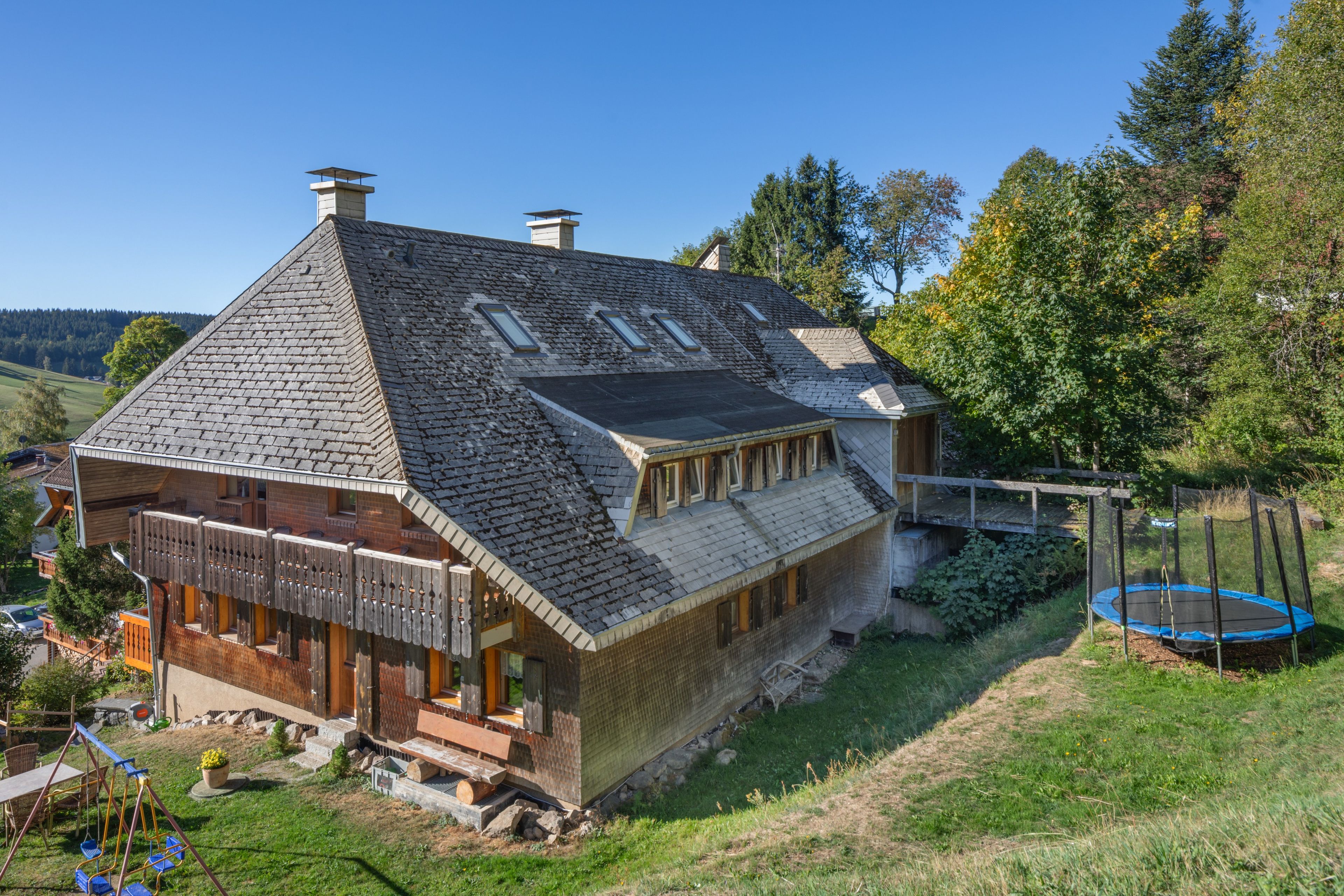 Une grande maison avec un toit en pente, de nombreuses cheminées et une cour clôturée avec un trampoline.