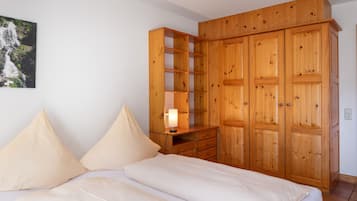 A bedroom with a bed, pillows, a wooden wardrobe, and a nightstand with a lamp.