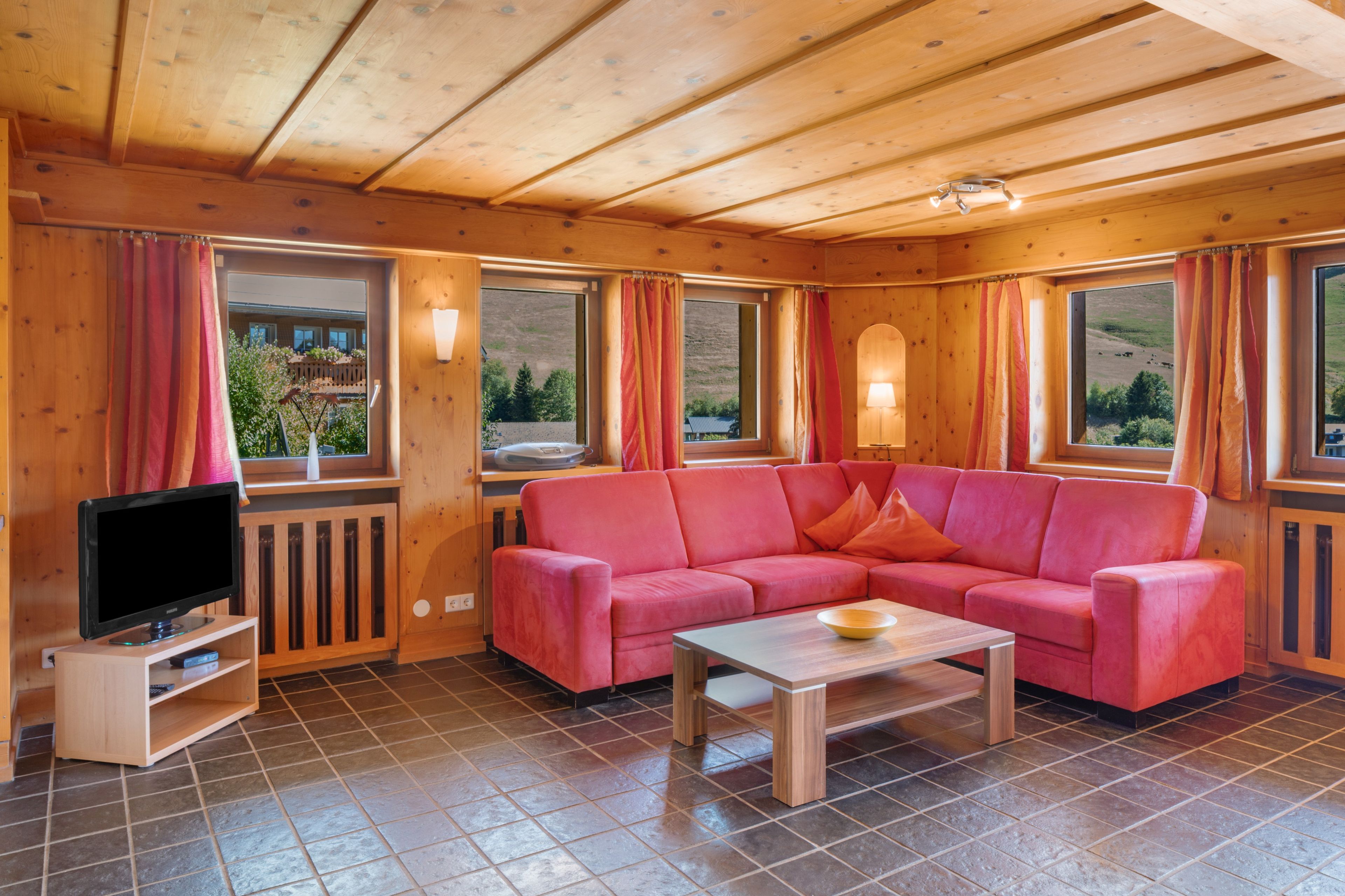 Ein gemütliches Zimmer mit Holzboden, einem roten Sofa, einem Fernseher auf einem Schrank und einem Fenster mit Vorhängen.