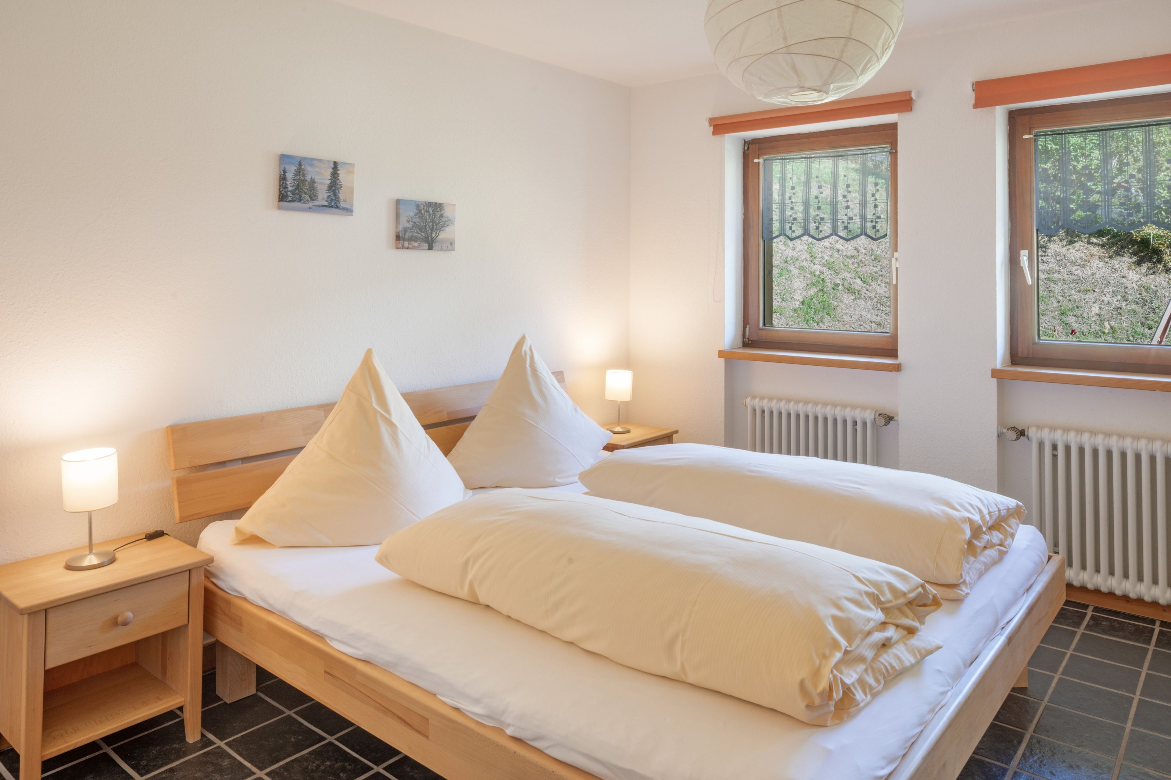 Un dormitorio con cama doble, dos mesitas de noche, un radiador y dos ventanas con cortinas.