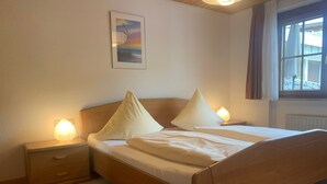 A bedroom with a wooden bed, two bedside tables with lamps, a nightstand, a window with curtains, and a painting on the wall. - Apartment 1 at Bühlhof in Todtnauberg (Todtnau)