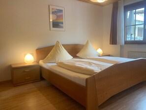 Ein Schlafzimmer mit einem hölzernen Bett, zwei Nachttischen mit Lampen, einem kleinen Tischchen, einem Fenster mit Vorhängen und einem Bild an der Wand.
