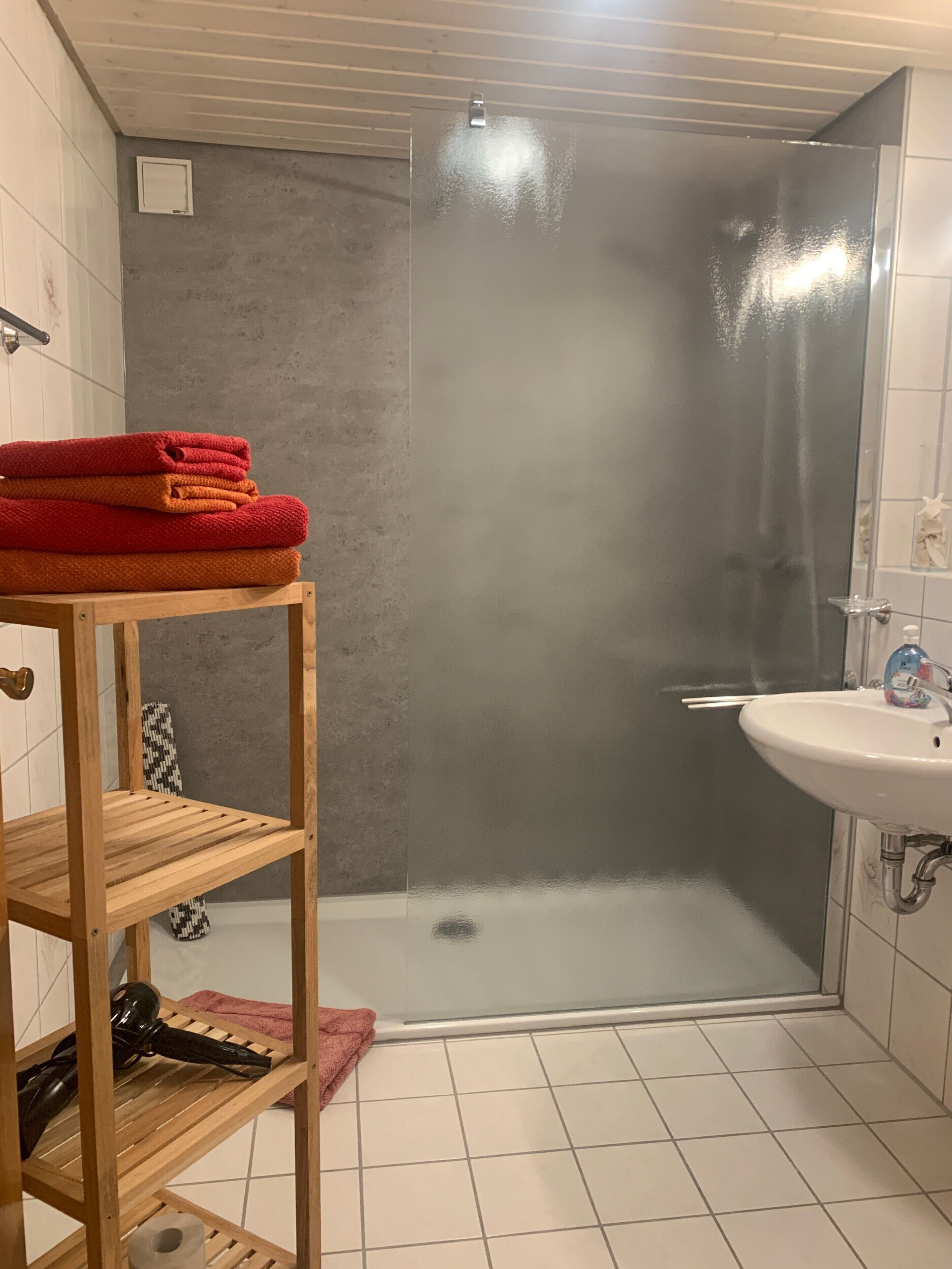 Une salle de bain avec une douche à parois de verre, une étagère en bois avec des serviettes, un lavabo et des toilettes.