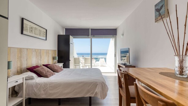 A hotel room with a bed, a desk, a chair, a television, and a view of the ocean. - Apartment in Las Palmas by Las Canteras Beach (Las Palmas de Gran Canaria)