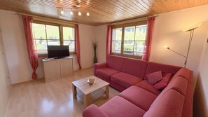 Ein Zimmer mit Holzboden, einem roten Sofa, einem Fernseher auf einem Schrank und großen Fenstern mit Vorhängen.