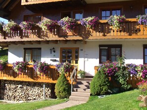 Ein zweistöckiges Gebäude mit einem hölzernen Balkon, auf der Tür ist die Nummer '20' sichtbar und die Balkonbrüstungen sind mit Blumenkästen versehen.